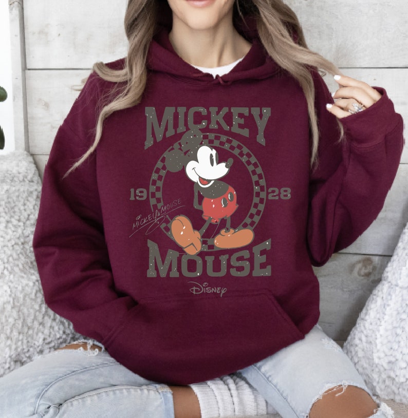 May include: Burgundy hoodie featuring a graphic of Mickey Mouse. The design includes the words "Mickey Mouse" and the years "1928" and "19". The Disney logo is at the bottom. The hoodie has a front pocket and a drawstring hood.