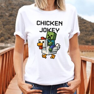 May include: White t-shirt with the text "CHICKEN JOKEY" in black block letters. A pixelated graphic shows a zombie riding a chicken. The chicken is white with a red comb and yellow feet. The zombie is green and blue.