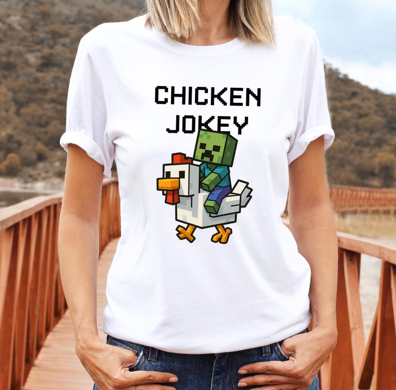 May include: White t-shirt with the text "CHICKEN JOKEY" in black block letters. A pixelated graphic shows a zombie riding a chicken. The chicken is white with a red comb and yellow feet. The zombie is green and blue.