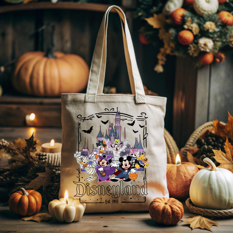 May include: Beige canvas tote bag featuring a Halloween-themed Disneyland design. The graphic includes cartoon characters in costumes, a castle, bats, and the Disneyland logo. Pumpkins and candles surround the bag.