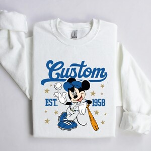May include: White sweatshirt featuring a cartoon Mickey Mouse in a baseball uniform, holding a bat and ball. The design includes the word "Custom" in blue script, the year "EST. 1958", and gold stars. The sweatshirt has long sleeves.