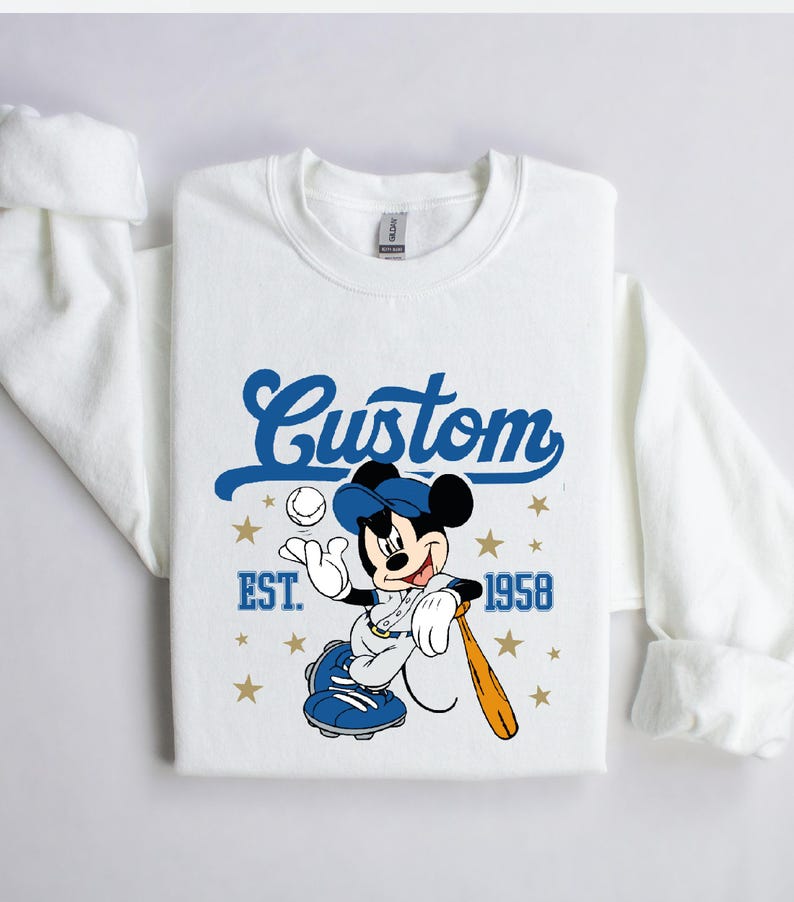 May include: White sweatshirt featuring a cartoon Mickey Mouse in a baseball uniform, holding a bat and ball. The design includes the word "Custom" in blue script, the year "EST. 1958", and gold stars. The sweatshirt has long sleeves.