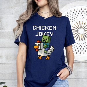 May include: A navy blue t-shirt featuring the text "CHICKEN JOKEY" above a pixelated graphic of a zombie riding a white chicken. The chicken has orange feet and a red comb. The zombie is green and blue.