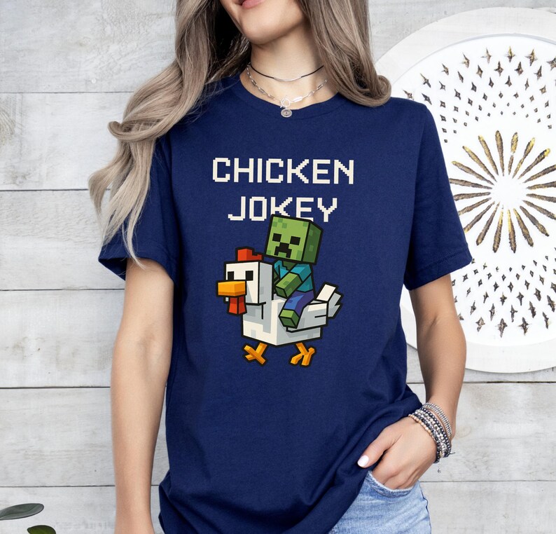 May include: A navy blue t-shirt featuring the text "CHICKEN JOKEY" above a pixelated graphic of a zombie riding a white chicken. The chicken has orange feet and a red comb. The zombie is green and blue.