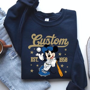 May include: Navy blue sweatshirt featuring a baseball-themed Mickey Mouse graphic. The design includes the word "Custom" in gold script, the year "1958", and stars. Mickey is depicted in a baseball uniform, holding a bat and ball.