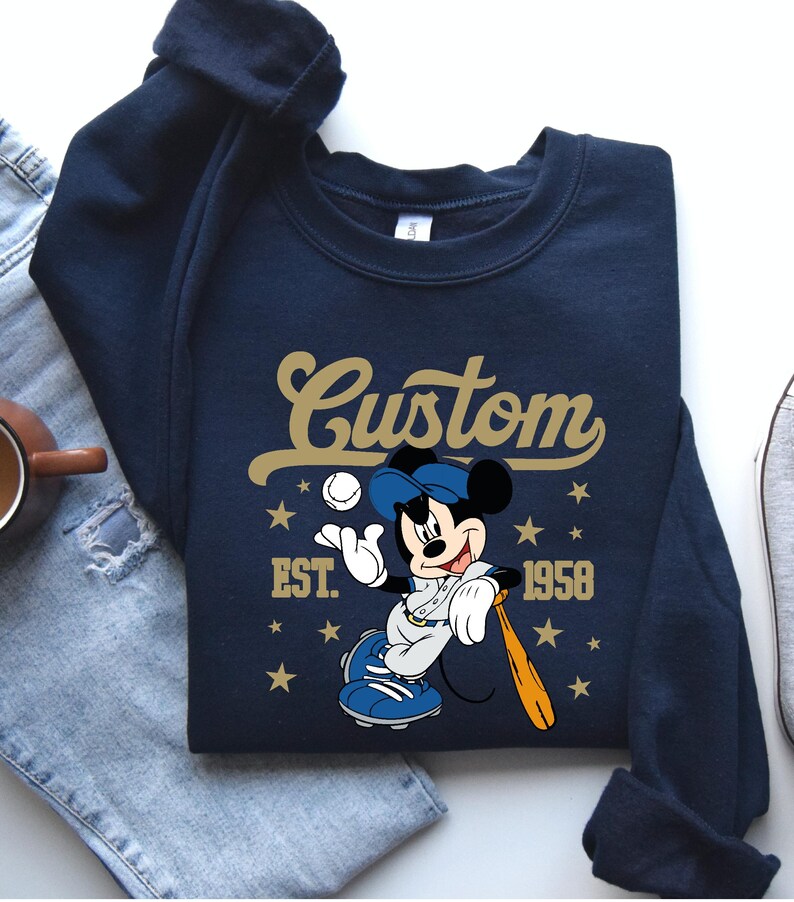 May include: Navy blue sweatshirt featuring a baseball-themed Mickey Mouse graphic. The design includes the word "Custom" in gold script, the year "1958", and stars. Mickey is depicted in a baseball uniform, holding a bat and ball.