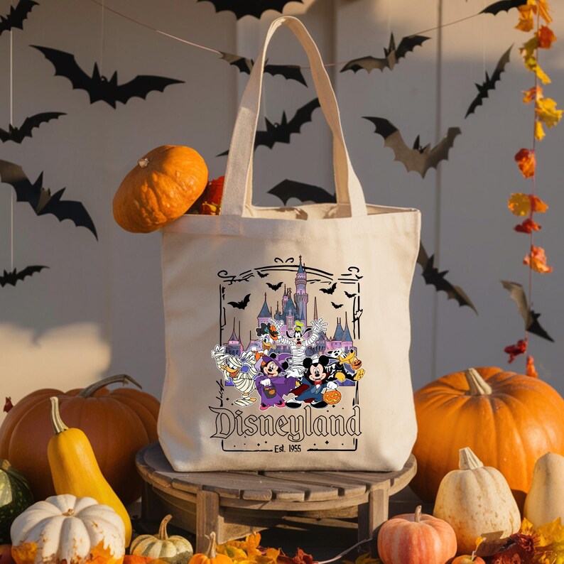 May include: A cream-colored tote bag featuring a Disneyland Halloween design with Mickey Mouse, Minnie Mouse, and other Disney characters in costume. The bag is surrounded by pumpkins and fall decorations.