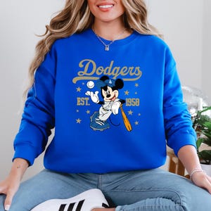 May include: Royal blue sweatshirt with a gold "Dodgers" script and a Mickey Mouse baseball design. The design includes stars, the text "EST. 1958", and a baseball bat. A classic sports-themed garment.