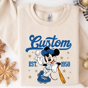 May include: Cream-colored sweatshirt featuring a baseball-themed Mickey Mouse graphic. The design includes the word "Custom" in blue script, the year "1958", and stars. Mickey is wearing a baseball uniform and holding a bat.
