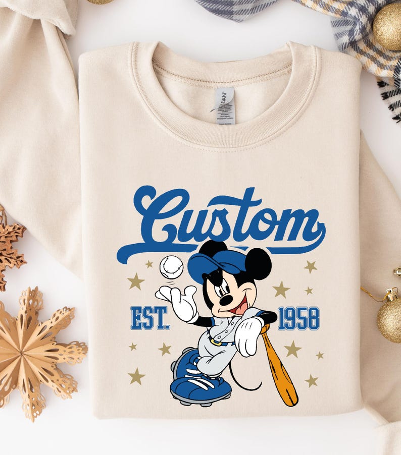 May include: Cream-colored sweatshirt featuring a baseball-themed Mickey Mouse graphic. The design includes the word "Custom" in blue script, the year "1958", and stars. Mickey is wearing a baseball uniform and holding a bat.