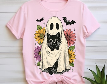May include: Pink t-shirt featuring a whimsical Halloween design. The graphic depicts a ghost holding a black cat, surrounded by colorful flowers and bats. The design is printed on a light pink t-shirt.