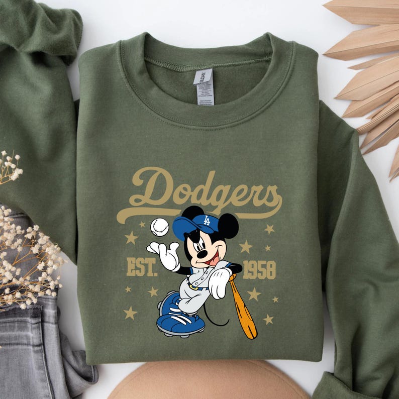 May include: An olive green sweatshirt with a Mickey Mouse baseball graphic. The design includes the word "Dodgers" in gold, Mickey Mouse in a baseball uniform, a baseball, and the text "EST. 1958". Gold stars are scattered around the design.