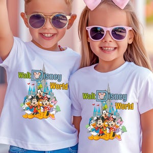 Puede incluir: Dos ni&ntilde;os con camisetas blancas con un gr&aacute;fico colorido de personajes de Disney y el texto "Walt Disney World". El gr&aacute;fico incluye a Mickey Mouse, Minnie Mouse, Pato Donald y Goofy. Ambos ni&ntilde;os llevan gafas de sol.