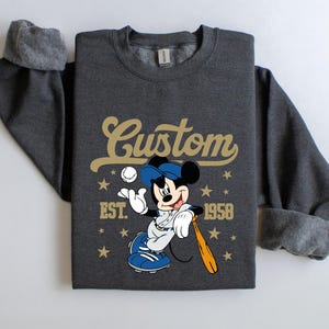 May include: Dark gray sweatshirt featuring a baseball-themed Mickey Mouse graphic. The design includes the word "Custom" in gold script, the year "1958", and stars. Mickey is depicted in a baseball uniform, holding a bat and ball.
