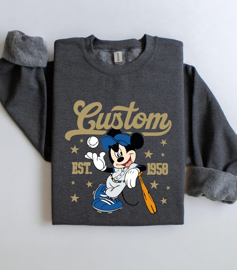 May include: Dark gray sweatshirt featuring a baseball-themed Mickey Mouse graphic. The design includes the word "Custom" in gold script, the year "1958", and stars. Mickey is depicted in a baseball uniform, holding a bat and ball.