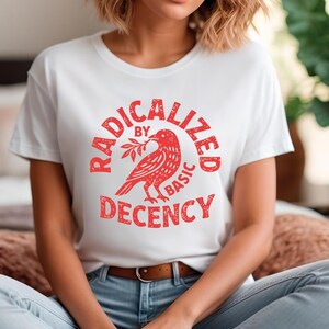 May include: White t-shirt with a red graphic. The graphic features a bird holding a branch, surrounded by the words "RADICALIZED BY BASIC DECENCY" in a circular design. The shirt is worn by a person.