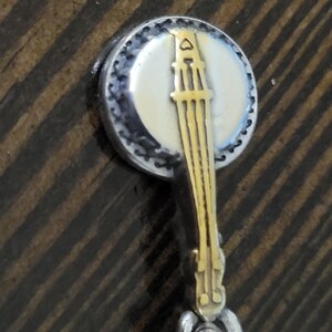 Puede incluir: Un dije en forma de banjo con un cuerpo de metal plateado y un dise&ntilde;o de banjo dorado. El dije tiene una cara blanca y crema con un borde moteado negro. El dije est&aacute; unido a un lazo de metal retorcido plateado.