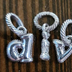 May include: Four silver charms with twisted bale loops. The charms include the letters "d", "v", and "a", and a small figure. The letters have heart cutouts and decorative details. The charms are on a wooden surface.