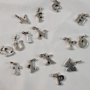 May include: A collection of silver-toned alphabet charms, each with a decorative swirl design. The charms are in various letter shapes, including 'V', 'F', 'G', 'H', 'N', 'U', 'I', 'Q', 'Y', 'O', 'T', 'L', 'X', 'A', and 'P'. Each charm has a small clasp.