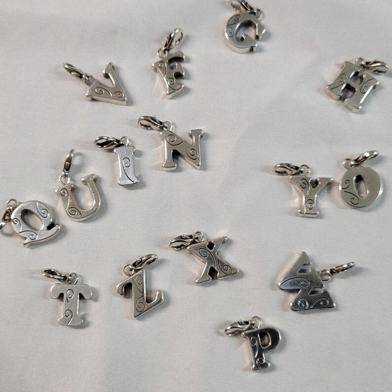 May include: A collection of silver-toned alphabet charms, each with a decorative swirl design. The charms are in various letter shapes, including 'V', 'F', 'G', 'H', 'N', 'U', 'I', 'Q', 'Y', 'O', 'T', 'L', 'X', 'A', and 'P'. Each charm has a small clasp.