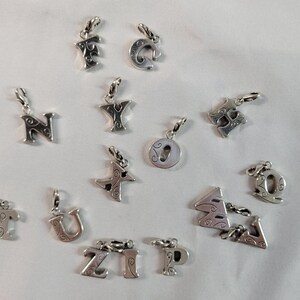 May include: A collection of silver-toned alphabet charms, each with a small loop for attachment. The letters include 'F', 'C', 'N', 'Y', 'T', 'U', 'Z', 'I', 'P', 'Q', and others. The charms are scattered on a white surface.