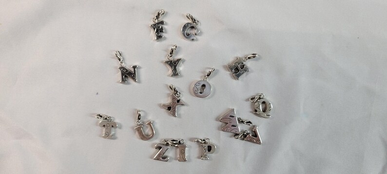 May include: A collection of silver-toned alphabet charms, each with a small loop for attachment. The letters include 'F', 'C', 'N', 'Y', 'T', 'U', 'Z', 'I', 'P', 'Q', and others. The charms are scattered on a white surface.