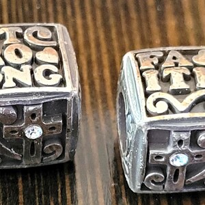 May include: Two silver-toned cube-shaped beads. One bead reads "STRONG" with a heart, the other reads "FAITH". Each bead features a cross with a small crystal and decorative swirls.