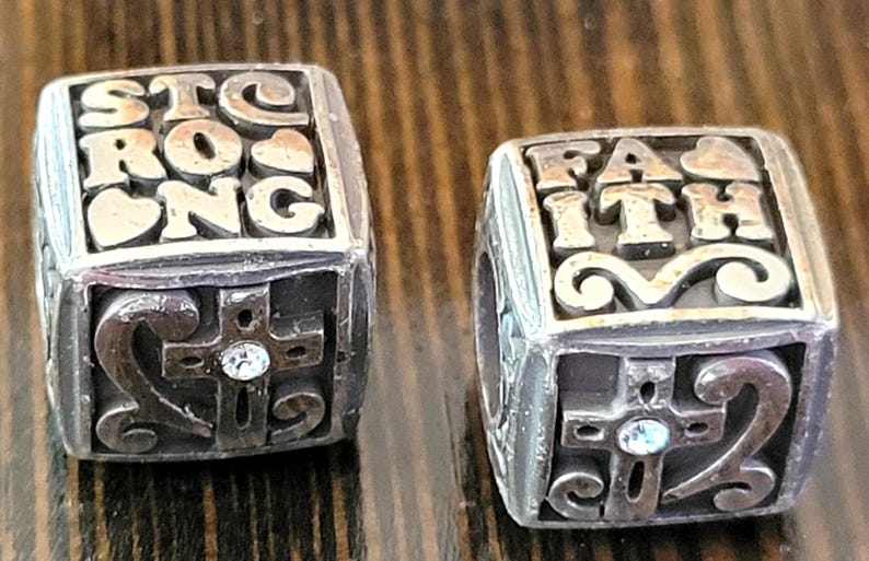 May include: Two silver-toned cube-shaped beads. One bead reads "STRONG" with a heart, the other reads "FAITH". Each bead features a cross with a small crystal and decorative swirls.