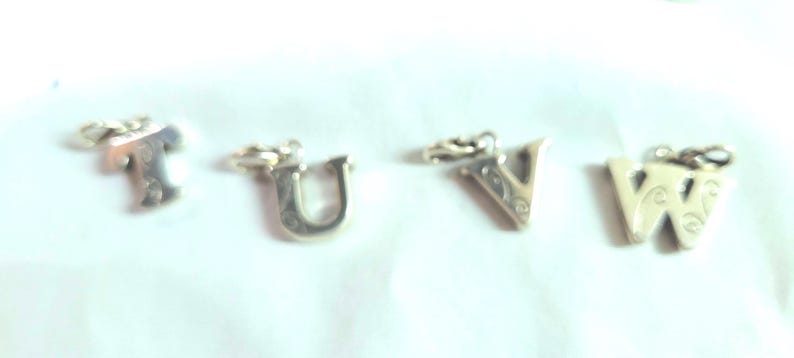 May include: Four silver-colored alphabet charms: T, U, V, and W. Each charm has a small loop at the top for attaching to a chain or bracelet. The letters have a decorative design.
