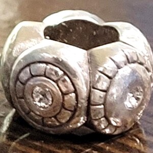 May include: A silver-toned, spherical bead with a geometric design. The bead features a central, circular pattern with a small, sparkling accent. The overall design is detailed and textured, with a polished finish.