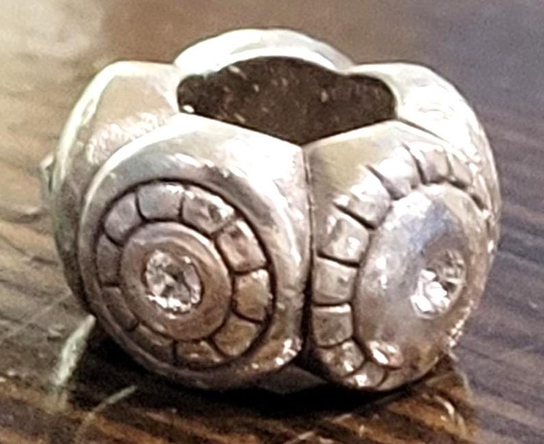 May include: A silver-toned, spherical bead with a geometric design. The bead features a central, circular pattern with a small, sparkling accent. The overall design is detailed and textured, with a polished finish.