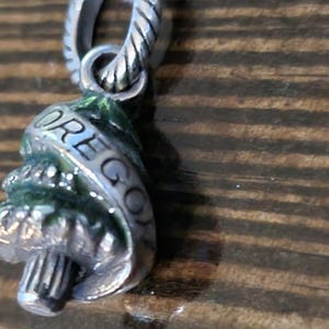 Puede incluir: Un charm de plata con la palabra "OREGON" grabada. El charm tiene forma de &aacute;rbol y tiene detalles de esmalte verde. El charm est&aacute; unido a un lazo de plata.