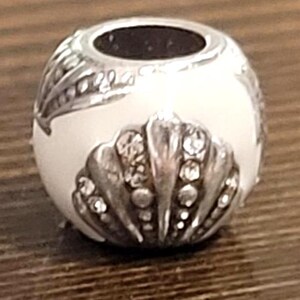 May include: A silver-toned bead with a white enamel finish. The bead features a seashell design with clear crystal accents and a leaf pattern. The top has a dark opening. This decorative bead is suitable for jewelry making.