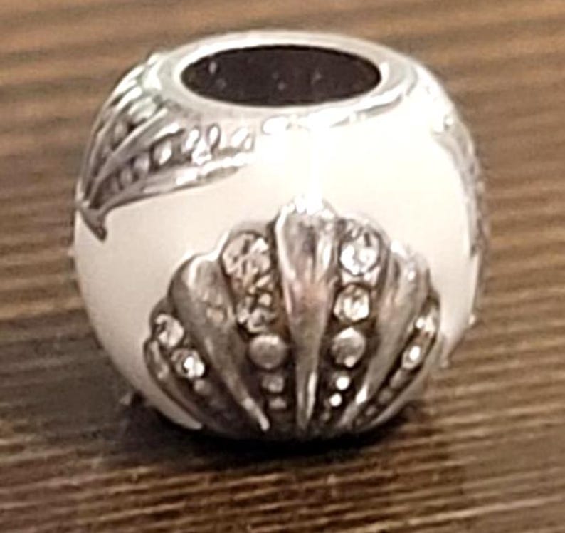 May include: A silver-toned bead with a white enamel finish. The bead features a seashell design with clear crystal accents and a leaf pattern. The top has a dark opening. This decorative bead is suitable for jewelry making.