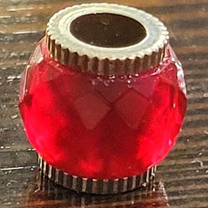May include: A faceted, red glass bottle stopper with a silver-colored metal rim and base. The stopper has a round shape and a decorative, ridged edge. The top opening is dark, contrasting with the bright red glass.
