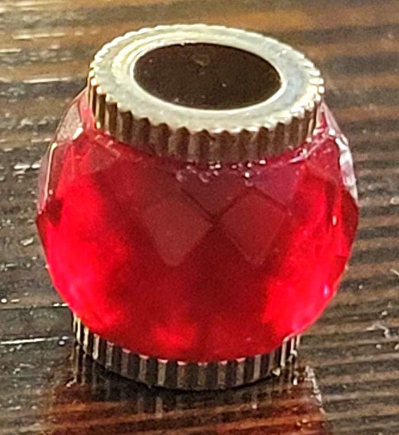 May include: A faceted, red glass bottle stopper with a silver-colored metal rim and base. The stopper has a round shape and a decorative, ridged edge. The top opening is dark, contrasting with the bright red glass.