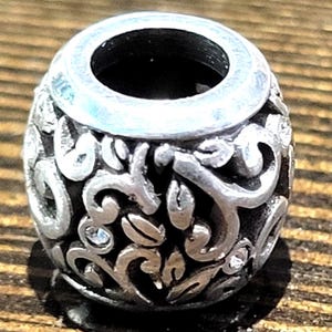 May include: A silver-colored bead with an open center and intricate floral and leaf patterns. The bead has a cylindrical shape with a slightly flared top. Small, clear crystals are interspersed within the design, adding a touch of sparkle.