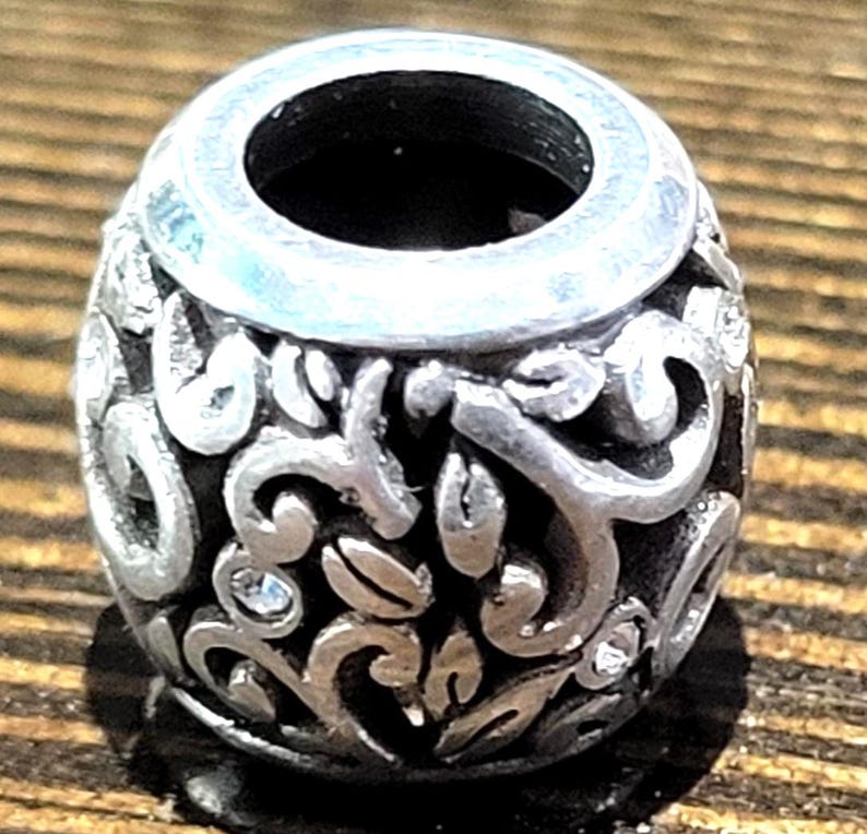 May include: A silver-colored bead with an open center and intricate floral and leaf patterns. The bead has a cylindrical shape with a slightly flared top. Small, clear crystals are interspersed within the design, adding a touch of sparkle.