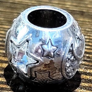 May include: A silver-toned charm bead with star and crystal accents. The bead has a large central hole and is decorated with various star shapes and small, clear crystals. The bead is round and has a polished finish.