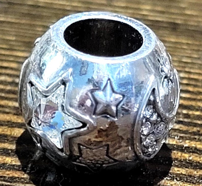 May include: A silver-toned charm bead with star and crystal accents. The bead has a large central hole and is decorated with various star shapes and small, clear crystals. The bead is round and has a polished finish.