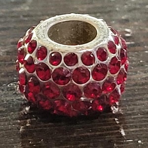 May include: A silver-toned bead with a cylindrical shape, encrusted with numerous round, ruby-red rhinestones. The bead has a central hole, and the rhinestones are closely set, creating a sparkling, textured surface. This decorative bead is suitable for jewelry making.