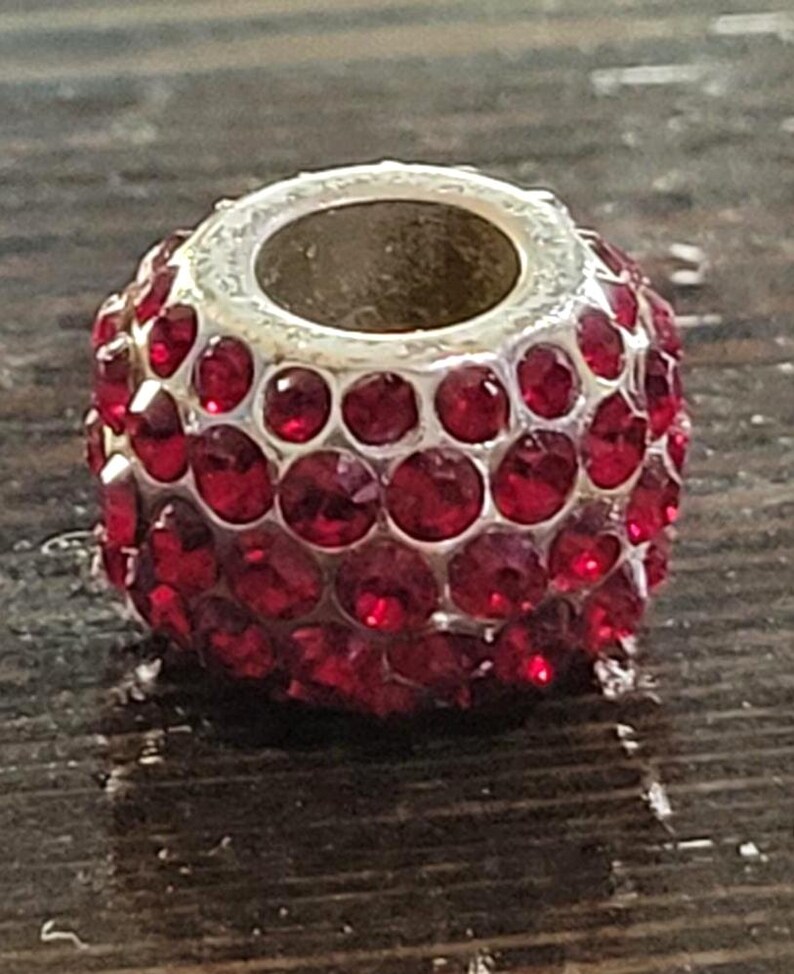 May include: A silver-toned bead with a cylindrical shape, encrusted with numerous round, ruby-red rhinestones. The bead has a central hole, and the rhinestones are closely set, creating a sparkling, textured surface. This decorative bead is suitable for jewelry making.