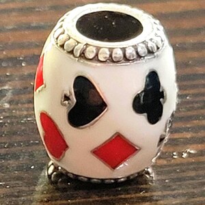 May include: A white bead with playing card suit symbols in red and black. The bead has a silver-colored top and bottom with a decorative pattern. The suits include hearts, diamonds, clubs, and spades.