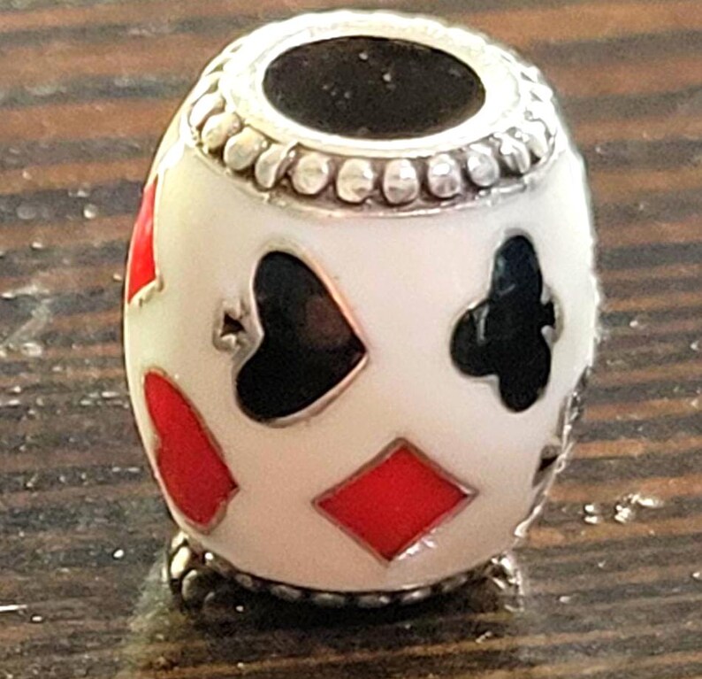 May include: A white bead with playing card suit symbols in red and black. The bead has a silver-colored top and bottom with a decorative pattern. The suits include hearts, diamonds, clubs, and spades.