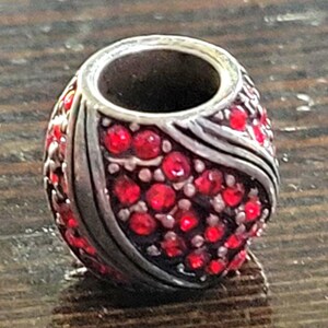 May include: A silver-toned bead with a central hole, covered in small, bright red gemstones. The bead has a rounded shape with a decorative silver band. The gemstones are closely set, creating a sparkling effect. This bead is suitable for jewelry making.