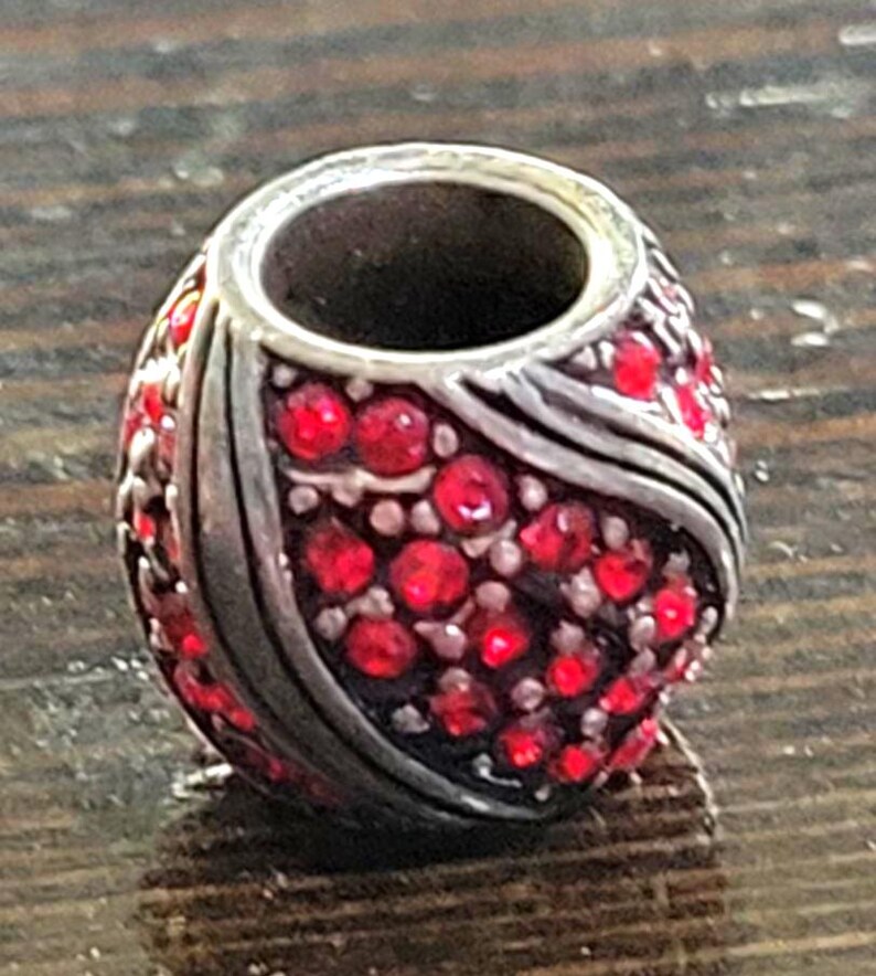 May include: A silver-toned bead with a central hole, covered in small, bright red gemstones. The bead has a rounded shape with a decorative silver band. The gemstones are closely set, creating a sparkling effect. This bead is suitable for jewelry making.
