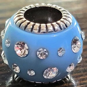 May include: A light blue bead with silver-colored accents and clear, sparkling rhinestones. The bead has a round shape with a silver-colored rim around the opening. Rhinestones of varying sizes are embedded in the blue surface.