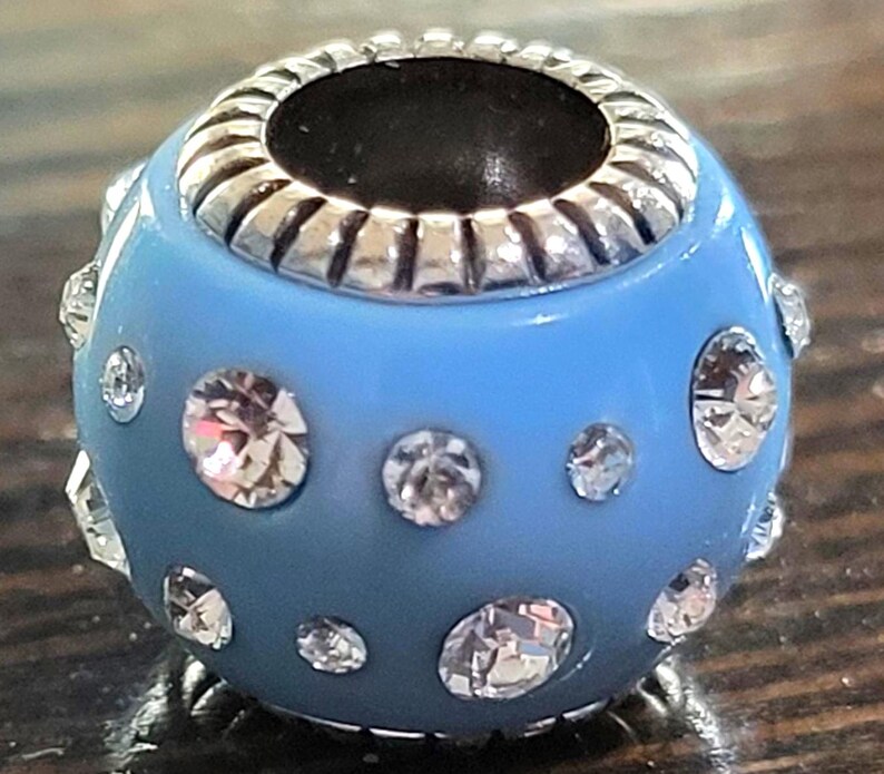 May include: A light blue bead with silver-colored accents and clear, sparkling rhinestones. The bead has a round shape with a silver-colored rim around the opening. Rhinestones of varying sizes are embedded in the blue surface.