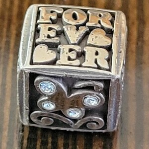 May include: Silver-toned charm with the word "FOREVER" spelled out in raised letters. The charm has a rectangular shape with a butterfly design and sparkling accents on one side. Two hearts are also visible.