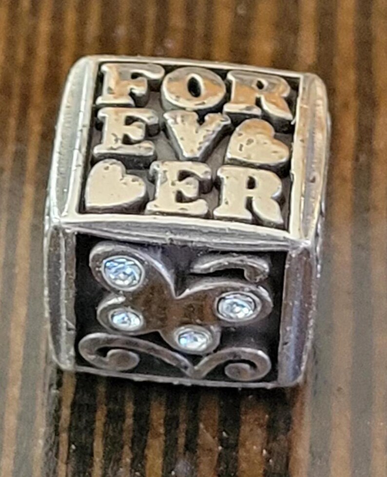 May include: Silver-toned charm with the word "FOREVER" spelled out in raised letters. The charm has a rectangular shape with a butterfly design and sparkling accents on one side. Two hearts are also visible.
